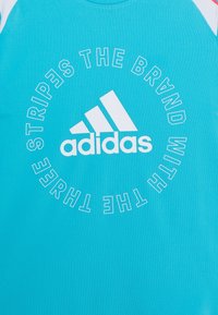 Camiseta deportiva turquesa con un logotipo circular blanco y la marca de tres rayas de Adidas. Presenta mangas cortas y una textura suave.