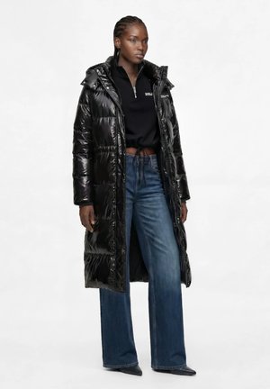 Kvinde med flettet hår iført en lang sort skinnende dunjakke, cropped sort lynlåstop, vidde blå jeans og sorte spidse sko.