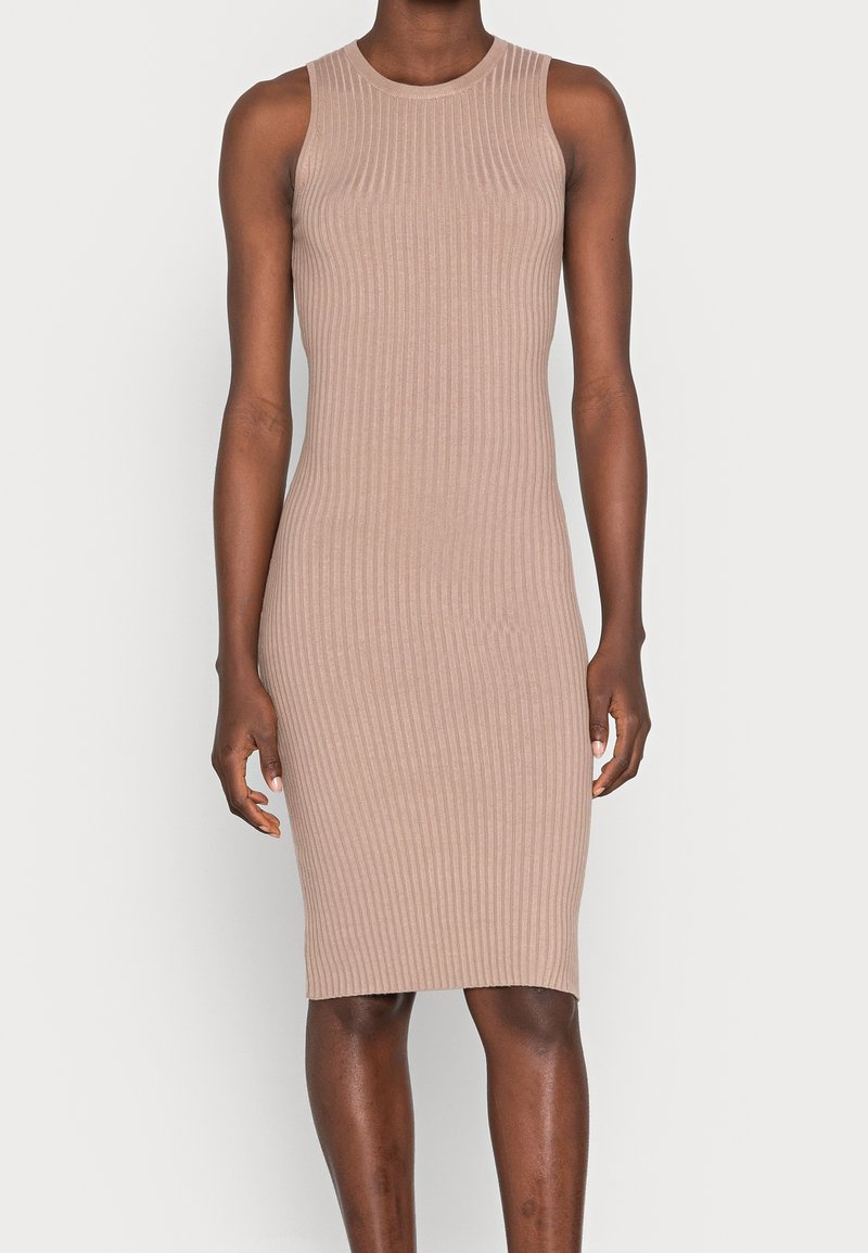 Robe beige en maille côtelée sans manches, longue jusqu'aux genoux, portée par une personne à la peau foncée, debout devant un fond clair uni.