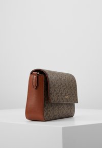 Hnědá kožená a vzorovaná krosbody taška s klopou, zlaté logo DKNY, vystavená na bílem podstavci na šedém pozadí.