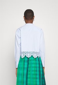 Vaaleansininen nappikauluspaita, jossa on valkoinen pitsireunus, yhdistettynä vihreään ruudulliseen hameeseen, jossa on tummia pystysuoria viivoja. Takaviisto.