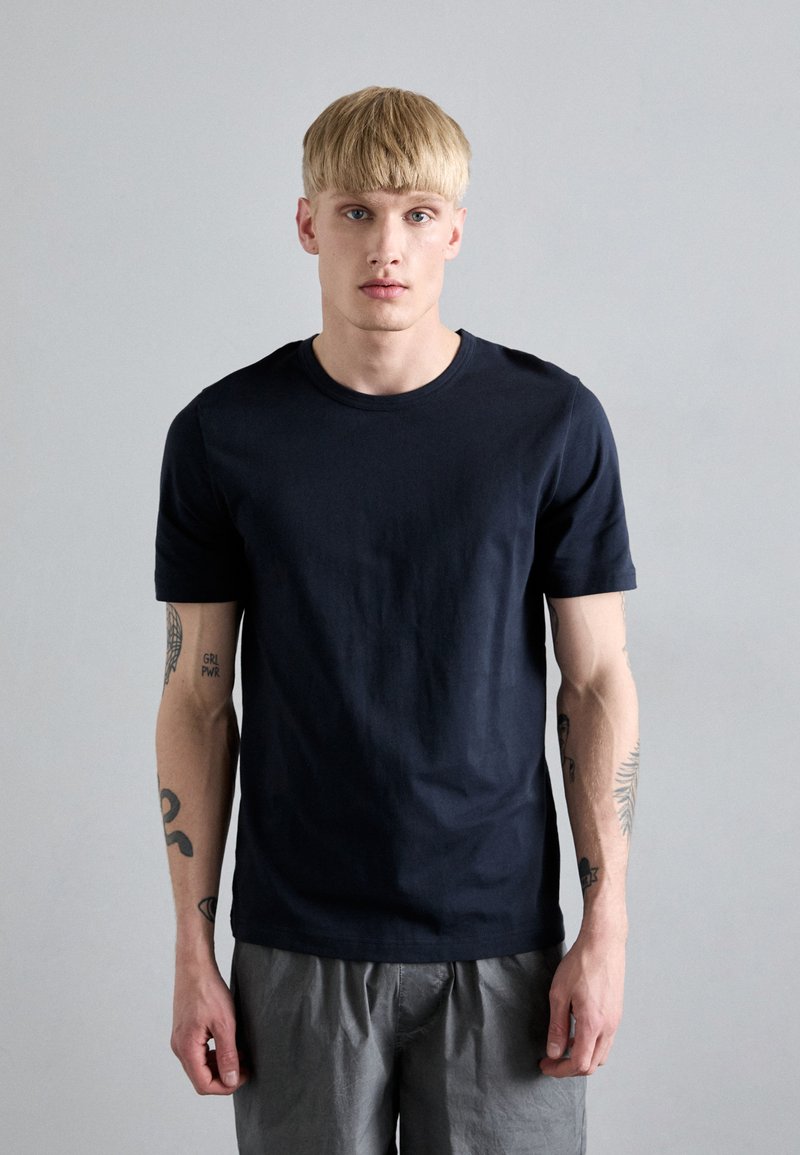 Marine crewneck t-skjorte i bomull med korte ermer; glatt tekstur; brukt med lysegrå shorts; tatoveringer synlige på armene.