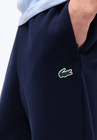 Άτομο που φοράει παντελόνι σε σκούρο μπλε με κεντημένο πράσινο λογότυπο κροκοδείλου κοντά στην τσέπη, με το χέρι να είναι μερικώς μέσα στην τσέπη.