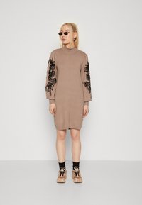 Strikkjole i beige med høj krave. Sort blomsterbroderi på oversized ærmer. Matcher med brune sneakers og sorte sokker.