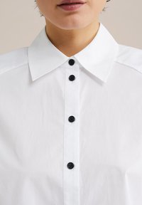 Camisa blanca de botones con un collar clásico, botones negros y una textura de tela suave, con una apariencia limpia y nítida.