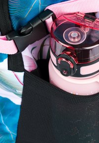 Růžová láhev na vodu s červeným víčkem je umístěna v černé síťované kapse batohu zdobeném barevnými květinovými vzory a růžovými popruhy.