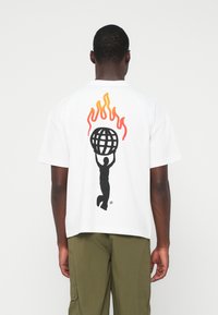 Homem vestindo uma t-shirt branca com uma silhueta preta segurando um globo acima da cabeça, com chamas laranja e vermelhas por trás.