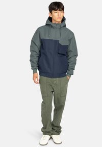 Giacca con cappuccio grigia e blu navy, con zip anteriore, polsini elasticizzati e tasca sul petto. Indossata con pantaloni verde oliva e sneakers bianche.