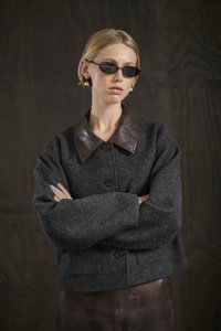 Une femme aux cheveux blonds courts porte des lunettes de soleil noires, des boucles d'oreilles en perles, une veste texturée gris foncé avec un col en cuir, et croise les bras.
