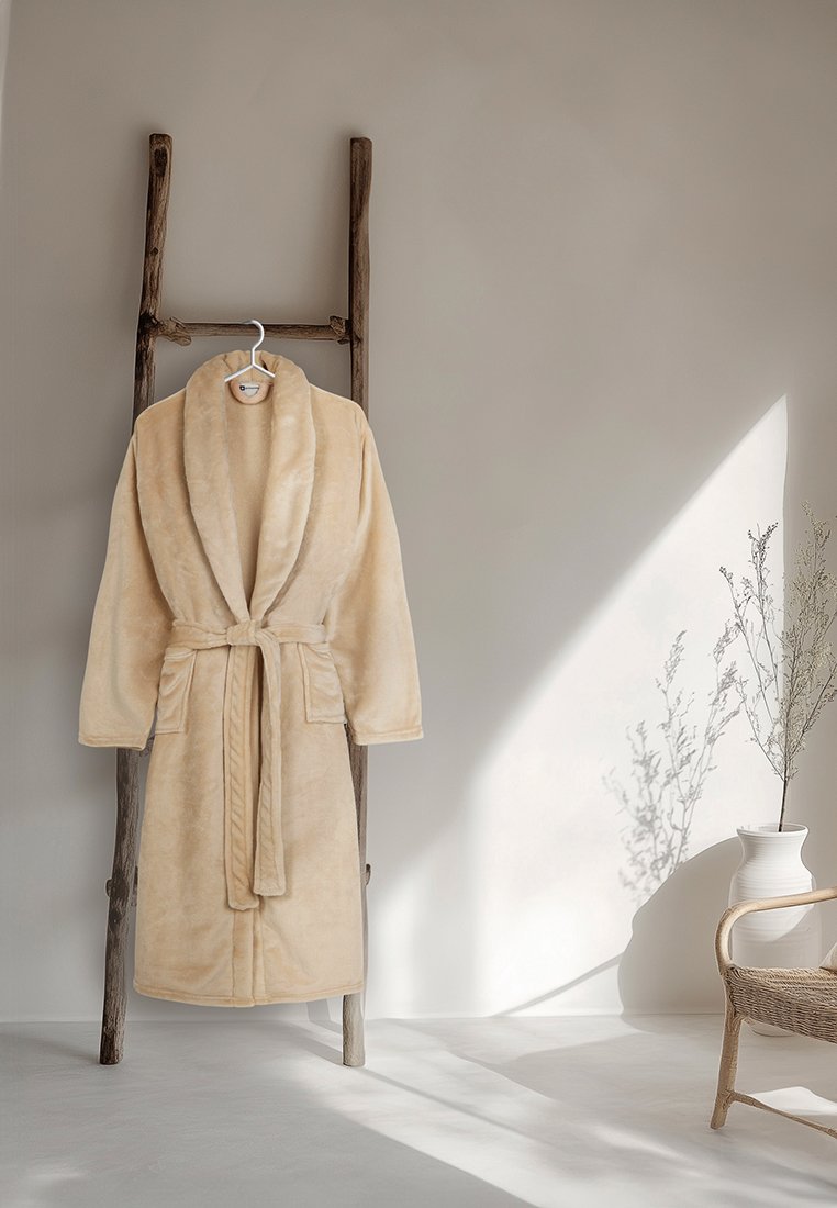 Beige, weicher Bademantel mit Schalkragen und Gürtel, der an einer Holzleiter vor einer neutralen Wand hängt; sanfte Textur und nahtloses Design.