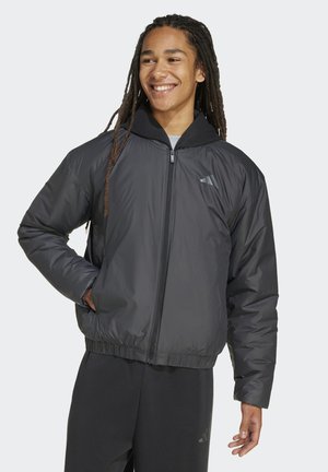 Persona sorridente con lunghi dreadlocks, indossa una giacca imbottita nera con cerniera e pantaloni neri, in piedi con una mano in tasca.