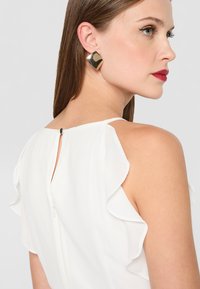 Bílý top s volánovými rukávy, s klíčovou zadní částí a malým zapínáním na knoflík. Model nosí čtvercové zlaté náušnice. Hladká textura.