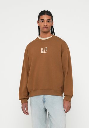Sudadera marrón de gran tamaño con el logo "GAP" en blanco, cuello redondo y puños acanalados, combinada con jeans de mezclilla claros.