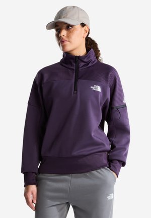Jeune personne aux cheveux bouclés portant une casquette beige clair, un pull violet à demi-fermeture éclair et un pantalon gris, debout avec une main dans la poche.