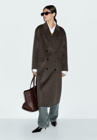 Manteau en laine marron avec un design à double boutonnage, associé à une chemise blanche, un accent orange, un jean bleu, des chaussures noires à bout pointu et un sac tote brun.