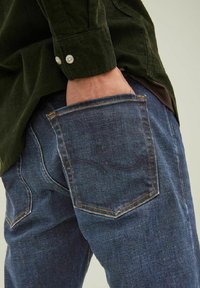 Jeans de mezclilla oscura con una forma ajustada, que presentan costuras en contraste, un bolsillo trasero y una camisa de pana verde texturizada en primer plano.