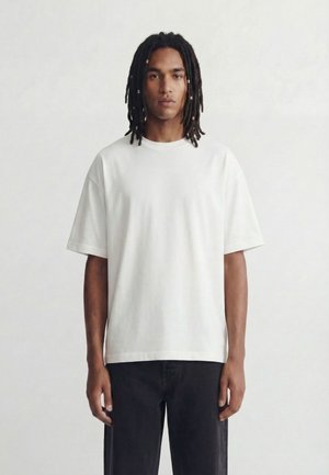 Young man with shoulder-length dreadlocks wearing a plain white oversized T-shirt and black pants, standing against a plain background.