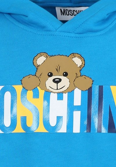 Sweatshirt bleu avec un graphique d'ours au visage souriant. Le texte "MOSCHINO" est imprimé en lettres majuscules en bleu et jaune.