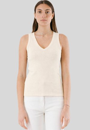 Donna con capelli castani lunghi, indossa un top crema senza maniche con scollo a V e pantaloni bianchi, in piedi davanti a uno sfondo grigio chiaro uniforme.