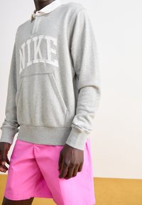 Szara bluza z kapturem z białym napisem „NIKE”, z przednią kieszenią i kołnierzykiem. Połączona z różowymi szortami, na neutralnym tle.