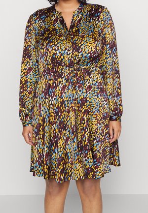 Femme portant une robe à manches longues, longueur genou, avec un motif abstrait coloré de points en jaune, bleu, blanc et bordeaux.