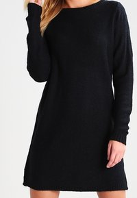 Svart stickad klänning med rund halsringning, lång ärm och avslappnad passform. Har ribbade manchetter och fåll. Mjuk struktur, minimalistiskt mönster.