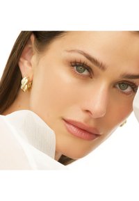 Orecchini a cerchio intrecciati in tono oro, finitura metallizzata, texture liscia, indossati da un modello con trucco neutro, che mette in evidenza le caratteristiche naturali.
