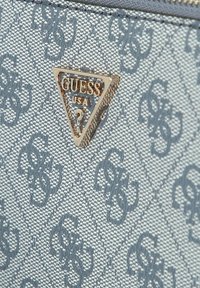 Logo triangolare in oro di Guess USA su tessuto grigio chiaro texture con motivo ripetuto di "GS" intrecciato, probabilmente per una borsa o un accessorio.