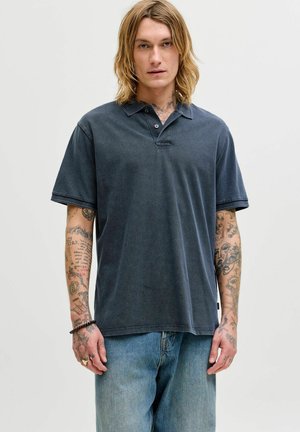 Ung person med langt blondt hår, tatoverede arme, iført en mørkegrå polo og blå jeans, stående mod en ensartet lys baggrund.