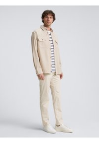 Beige longsleeve shirt met twee borstzakken, gedragen over een wit en marineblauw gestreept T-shirt. Lichtbeige broek en witte sneakers.