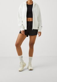 Sudadera blanca con cremallera, top corto negro, pantalones cortos negros, calcetines blancos con logo negro y zapatillas blancas con suelas texturizadas. Ropa atlética casual.