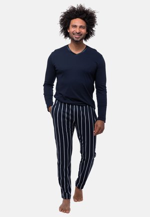 Uomo con capelli ricci che sorride, cammina a piedi nudi, indossa una camicia scura a maniche lunghe e pantaloni neri con strisce verticali bianche.