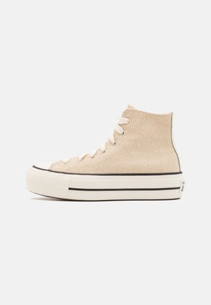 Des baskets montantes en toile texturée beige, avec des lacets blancs, une semelle en caoutchouc noire et un logo signature au talon arrière.