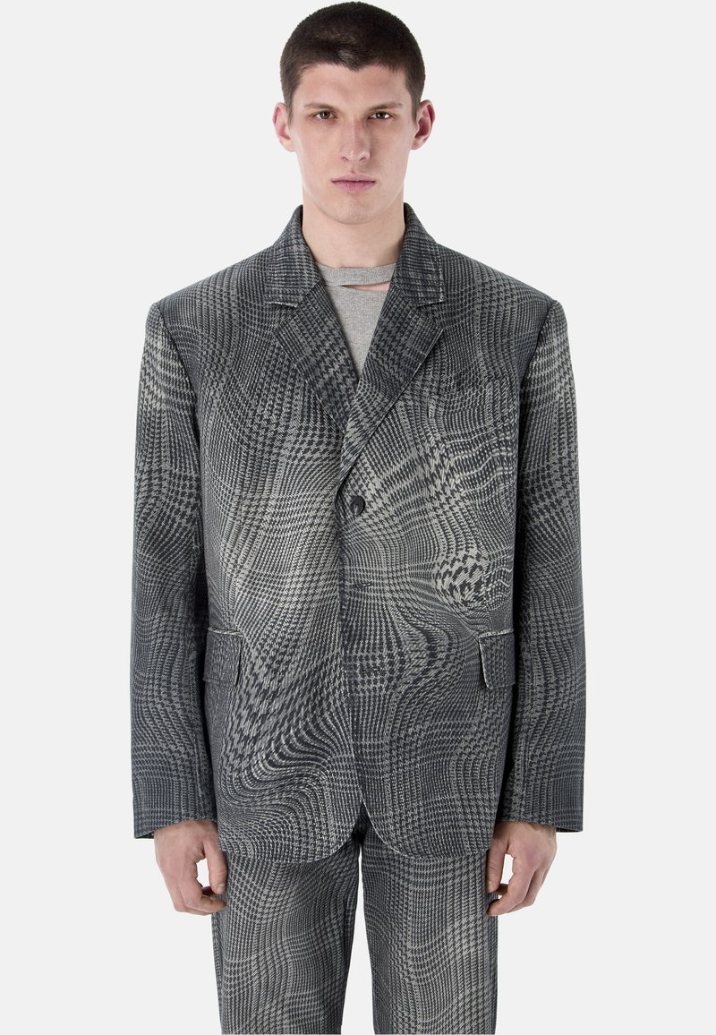 Joven con chaqueta de traje de cuadros gris grande y pantalones a juego, con camisa gris claro debajo, de pie frente a un fondo blanco.