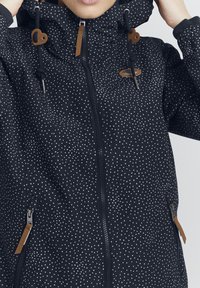 Giacca nera con pois bianchi, dotata di chiusura con zip, cappuccio regolabile con dettagli marroni e tasche laterali con zip.