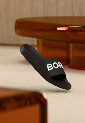 Čierna šľapka na platforme s bielym textom "BOR" viditeľným čiastočne, položená na béžovom koberci vedľa okrajov stolíka medenej priehľadnej farby.