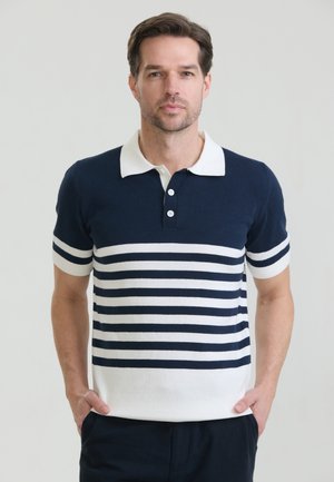 Man met kort bruin haar, draagend een marineblauw-wit gestreept poloshirt, staand met handen in de zakken tegen een lichte achtergrond.