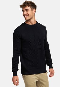 Zwarte trui met een gestructureerd chevronpatroon, ronde hals en geribde boorden. Gecombineerd met lichtgekleurde broek.