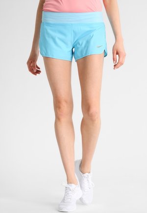 Kvinde iført lyseblå Nike-løbershorts, lyserød top og hvide sneakers, stående mod en ensfarvet hvid baggrund.