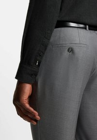 Camicia nera con bottoni realizzata in un tessuto testurizzato, abbinata a pantaloni grigi con un motivo sottile, tasca laterale e un singolo bottone sul retro.