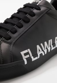 Sneaker in pelle nera con lacci neri e testo bianco "FLAWLESS" sul lato e sulla suola, primo piano dei dettagli anteriori e laterali.