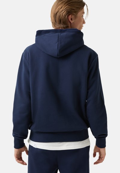 Osoba oblečená v navy modré mikině s kapucí a odpovídajících kalhotách, otočená zády, s bílým tričkem vykukujícím zpod mikiny.