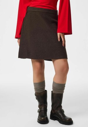 Femme portant une jupe trapèze marron foncé, des chaussettes grises froissées, des bottes noires mi-mollet et un haut rouge à manches longues évasées.