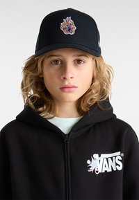 Gorra negra con diseño de llamas bordadas que presenta acentos en púrpura y naranja, usada por un niño con una sudadera con cremallera negra que tiene un logo.