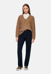 Brauner, strukturierter, übergroßer Pullover mit V-Ausschnitt, kombiniert mit dunkelblauen geraden Jeans und gemusterten Turnschuhen.