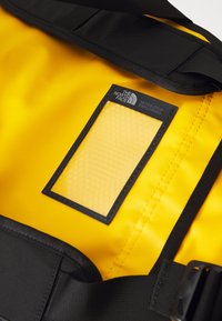 Bolsa impermeable amarilla con una correa negra, etiqueta de bolsillo de malla hexagonal y el logo de The North Face, con una textura lisa.
