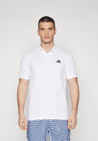 Camisa polo blanca de manga corta, con un logo de Adidas en negro y paneles laterales texturizados. Diseño de cuello alto y textura de tela suave.