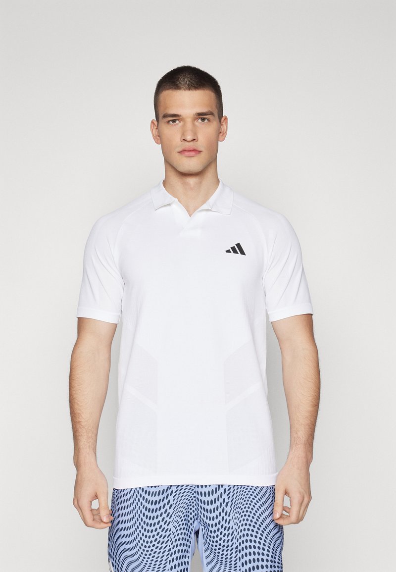 Camisa polo blanca de manga corta, con un logo de Adidas en negro y paneles laterales texturizados. Diseño de cuello alto y textura de tela suave.