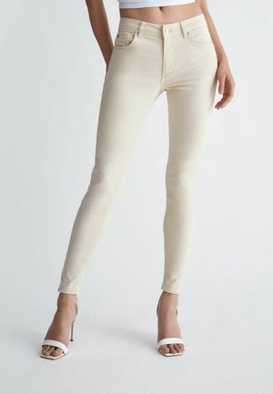 Vrouw met lichtbeige skinny jeans met hoge taille en witte sandalen met hak, staand tegen een effen achtergrond. Bovenlichaam niet zichtbaar.