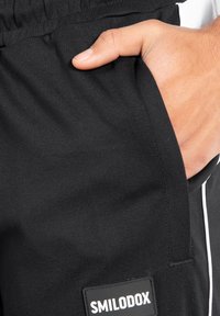 Svarta sweatpants har en sidoficka och en märkesetikett. Materialet är slätt med en lätt lyster, och det finns ett kontrasterande vitt streck.